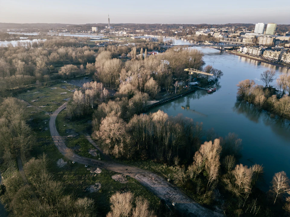 The image presents an aerial view of a river flowing through a city, surrounded by lush green trees and buildings on both sides. The river is calm with no visible boats or people present. On one side of the river, there are several tall buildings, including a large tower that stands out against the sky. The other side of the river features more residential areas with houses nestled among the trees.