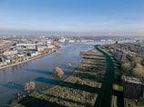 An aerial view of a river flowing through an urban landscape with a combination of industrial and residential buildings. Wind turbines are visible in the distance, along with lush greenery lining the riverbanks. The scene is clear and bright with a blue sky.