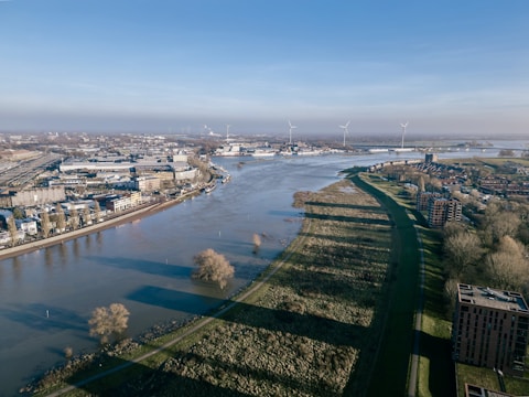 An aerial view of a river flowing through an urban landscape with a combination of industrial and residential buildings. Wind turbines are visible in the distance, along with lush greenery lining the riverbanks. The scene is clear and bright with a blue sky.