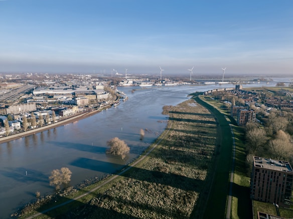 An aerial view of a river flowing through an urban landscape with a combination of industrial and residential buildings. Wind turbines are visible in the distance, along with lush greenery lining the riverbanks. The scene is clear and bright with a blue sky.