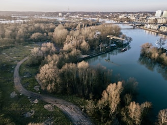 An aerial view of a serene landscape featuring a river surrounded by patches of trees. Buildings and infrastructure are evident in the background, along with a bridge crossing the water. The land is a mix of grass and pathways, with sparse tree coverage and a generally open area.