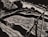 A monochromatic illustration featuring a dynamic composition with curved and sharp lines. It includes abstract forms that resemble a river or stream flowing past angular and curved structures, possibly rocks or hills, with a prominent object resembling a wooden bridge or structure.