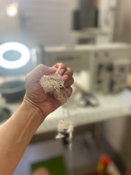 A hand holds a tangled bundle of threads in the foreground, with a sewing machine blurred in the background. The image suggests a focus on sewing or crafting, portraying an intimate view of textile work in progress.