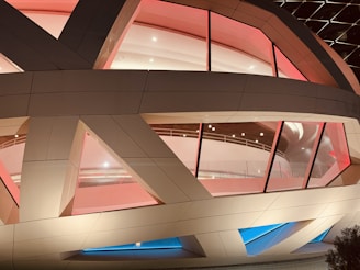 A modern architectural structure with large glass panels and geometric shapes. The building features warm lighting with hues of red and blue, creating a striking contrast. The interior appears to be spacious and well-lit with visible ceiling lights and a curved interior design.