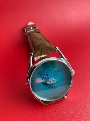 A watch with a brown leather strap is positioned against a red background. The watch face displays a whimsical illustration of a UFO abducting a cow, set against a blue backdrop.