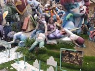 A vibrant and colorful sculpture display featuring fantastical figures in various poses. The scene includes human-like figures with exaggerated expressions and gestures set amidst lush greenery and decorative elements. A variety of textures and dynamic interactions among the characters create a sense of chaos or drama.