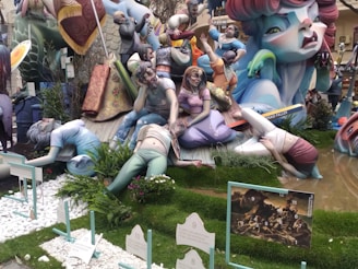 A vibrant and colorful sculpture display featuring fantastical figures in various poses. The scene includes human-like figures with exaggerated expressions and gestures set amidst lush greenery and decorative elements. A variety of textures and dynamic interactions among the characters create a sense of chaos or drama.