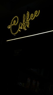 A brightly lit, stylized script sign spells out the word 'coffee' in yellow lights, creating a warm and inviting atmosphere. Below the sign, there are a few beverage cartons displayed in dim lighting.