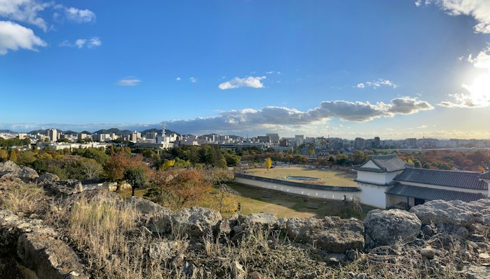 The image displays a panoramic view of a cityscape with an expansive sky overhead. In the foreground, there are rugged rocks and shrubbery, leading to a traditional Japanese castle structure with white walls and a dark roof. Beyond the castle lies a mix of modern city buildings and green, wooded areas. The sky is mostly clear with a few scattered clouds and the sun is shining brightly, casting shadows on the landscape.