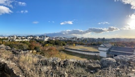 The image displays a panoramic view of a cityscape with an expansive sky overhead. In the foreground, there are rugged rocks and shrubbery, leading to a traditional Japanese castle structure with white walls and a dark roof. Beyond the castle lies a mix of modern city buildings and green, wooded areas. The sky is mostly clear with a few scattered clouds and the sun is shining brightly, casting shadows on the landscape.