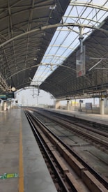 The image features a modern railway station platform with empty tracks and a high, arched roof. Metal beams and structural elements are visible, along with signs and lighting fixtures hanging from the ceiling. The platform is clean and wide, extending towards the horizon with clear skies visible through the transparent sections of the roof.