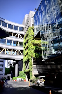 A modern architectural structure featuring a combination of glass and concrete. There are green plants growing vertically on one section of the building. The bright sunlight enhances the contrast between the greenery and the sleek design. Vehicles and a few people can be seen at the ground level, adding life to the scene.