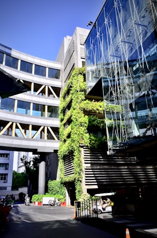 A modern architectural structure featuring a combination of glass and concrete. There are green plants growing vertically on one section of the building. The bright sunlight enhances the contrast between the greenery and the sleek design. Vehicles and a few people can be seen at the ground level, adding life to the scene.