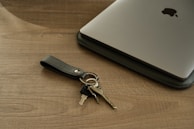 A sleek silver laptop lies on a wooden surface next to a black leather keychain holding several keys. The table has a smooth, natural wood grain that complements the metallic sheen of the laptop.