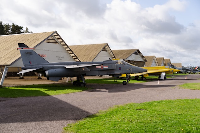 Two aircraft are displayed outdoors near a row of large, uniquely shaped hangars. The first aircraft is gray, parked on a paved surface, while the second is yellow. In the background, several other aircraft can be seen near similarly styled hangars. The sky is partly cloudy, and the surrounding area consists of grass and trees.