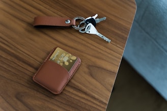 A wooden surface features a brown leather keychain holding a set of keys next to a brown card holder with a card partially visible inside.