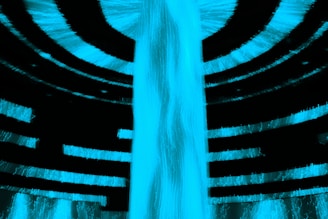 A bright, electric blue light forms a cascade that resembles a waterfall at the center, surrounded by dark, curved, overhanging layers creating an abstract and futuristic appearance. The blue glow is intense, standing out against the black background, suggesting a sense of movement and fluidity.