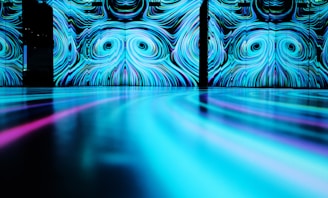 A large-scale digital art installation featuring swirling patterns of blue and cyan shades on a wall. The reflective surface in the foreground extends the patterns, creating a mesmerizing and immersive visual experience.
