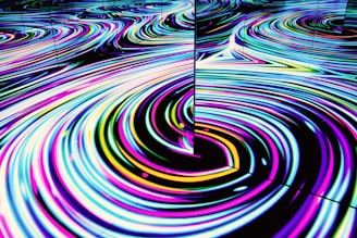 Vivid, swirling patterns of neon colors create a mesmerizing wave-like effect on a reflective surface. The movement is fluid and hypnotic, featuring loops and curves in an abstract form.