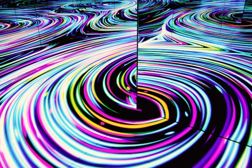 Vivid, swirling patterns of neon colors create a mesmerizing wave-like effect on a reflective surface. The movement is fluid and hypnotic, featuring loops and curves in an abstract form.
