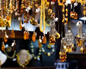 An array of amber jewelry, including necklaces and pendants, is displayed hanging in a showcase. The pieces are intricately designed with a range of warm amber tones, complemented by silver elements. Price tags are attached to each item, indicating they are for sale.