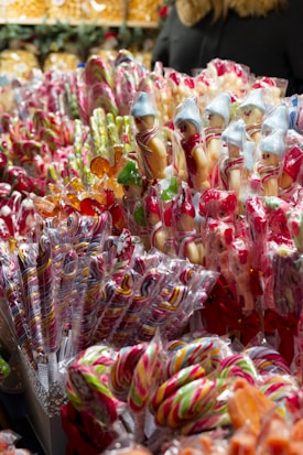 An array of colorful lollipops and candy sticks arranged in a market stall, featuring a variety of shapes and colors. Many of the candies are decorated with holiday-themed designs, such as small figures in festive attire.