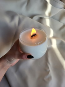 A hand is holding a white candle with a wooden wick that is lit, creating a warm flame. The background consists of soft, light-colored fabric that appears to be gently draped, adding a calm and cozy feel to the scene.