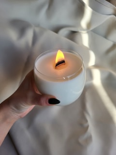 A hand is holding a white candle with a wooden wick that is lit, creating a warm flame. The background consists of soft, light-colored fabric that appears to be gently draped, adding a calm and cozy feel to the scene.