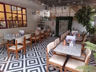 An indoor cafe with neatly arranged tables and chairs. Each table is set with copper water jugs, condiments, and menus. The decor includes greenery and a patterned black-and-white floor. Large windows with red frames add to the ambiance, and plants are strategically placed throughout the area.