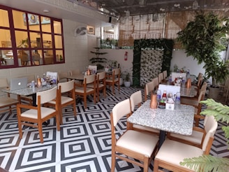 An indoor cafe with neatly arranged tables and chairs. Each table is set with copper water jugs, condiments, and menus. The decor includes greenery and a patterned black-and-white floor. Large windows with red frames add to the ambiance, and plants are strategically placed throughout the area.