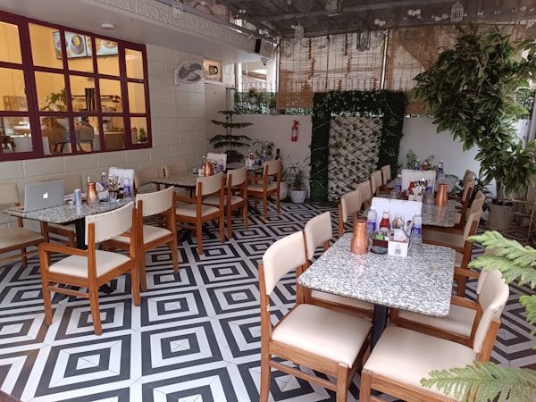 An indoor cafe with neatly arranged tables and chairs. Each table is set with copper water jugs, condiments, and menus. The decor includes greenery and a patterned black-and-white floor. Large windows with red frames add to the ambiance, and plants are strategically placed throughout the area.