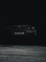 A dark, shadowy image featuring the rear view of a sports car with a license plate displaying 000000. The car is parked in a dimly lit area.