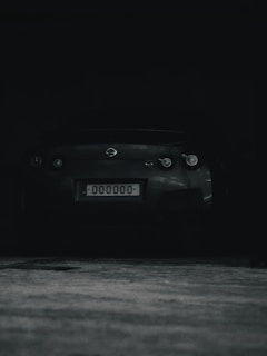 A dark, shadowy image featuring the rear view of a sports car with a license plate displaying 000000. The car is parked in a dimly lit area.