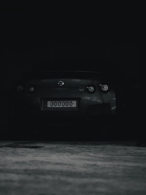 A dark, shadowy image featuring the rear view of a sports car with a license plate displaying 000000. The car is parked in a dimly lit area.