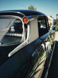 A close-up view of a vintage car with a sleek, glossy black paint job. The side mirrors and chrome detailing are prominent, reflecting the surroundings. The windows are slightly darkened, adding to its classic appeal. In the background, there are hints of greenery and outdoor elements, suggesting an outdoor setting possibly in a garden or park.