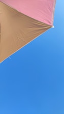 A triangular section of a beach umbrella in shades of tan and pink is set against a clear blue sky.