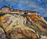 A landscape painting depicting a cluster of buildings situated on a rugged cliff overlooking the sea. The sky is filled with swirling clouds in blue and white, contrasting with the earthy tones of the cliff. The buildings appear to be rustic, with colors ranging from earthy reds to muted whites and browns, blending into the natural rock formations.
