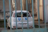 A white Fiat car marked with blue 'U N' letters is parked in an enclosed area. The car appears to be behind a blue fence with bars, and there is some greenery on the ground. The background includes chain-link fencing and a concrete wall.