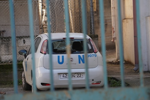 A white Fiat car marked with blue 'U N' letters is parked in an enclosed area. The car appears to be behind a blue fence with bars, and there is some greenery on the ground. The background includes chain-link fencing and a concrete wall.