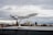 A model airplane is mounted on a stand with a cloudy city skyline in the background. The plane appears to be a detailed scale replica, prominently displaying the Lufthansa logo on the fuselage. The scene is overcast with muted colors, suggesting a calm atmosphere.