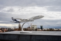 A model airplane is mounted on a stand with a cloudy city skyline in the background. The plane appears to be a detailed scale replica, prominently displaying the Lufthansa logo on the fuselage. The scene is overcast with muted colors, suggesting a calm atmosphere.