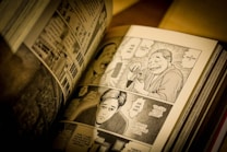 The image depicts an open manga book with black and white illustrations. The visible pages contain several panels showing various characters engaged in conversation, with speech bubbles conveying dialogue. The artwork is detailed, capturing expressions and actions of the characters.