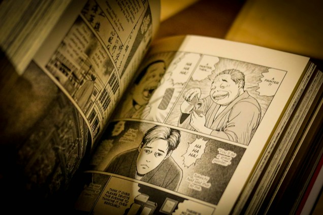 The image depicts an open manga book with black and white illustrations. The visible pages contain several panels showing various characters engaged in conversation, with speech bubbles conveying dialogue. The artwork is detailed, capturing expressions and actions of the characters.