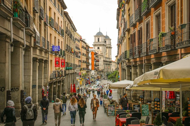 A bustling urban street lined with historic European architecture. Buildings have balconies with wrought iron railings, and the street is filled with pedestrians walking in various directions. Sidewalk cafes with umbrellas and outdoor seating areas populate the scene, with a prominent church or cathedral visible in the background. The overall atmosphere is lively and vibrant.
