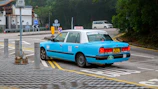 A light blue taxi is parked on the side of a road, near a sign that reads 'Alighting Only'. The vehicle has a yellow license plate with the number KY 8292, and the word 'TAXI' is displayed on its roof. There are several road signs and a small grey car in the background, indicating a quiet urban setting with surrounding greenery.