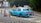 A light blue taxi is parked on the side of a road, near a sign that reads 'Alighting Only'. The vehicle has a yellow license plate with the number KY 8292, and the word 'TAXI' is displayed on its roof. There are several road signs and a small grey car in the background, indicating a quiet urban setting with surrounding greenery.