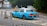 A light blue taxi is parked on the side of a road, near a sign that reads 'Alighting Only'. The vehicle has a yellow license plate with the number KY 8292, and the word 'TAXI' is displayed on its roof. There are several road signs and a small grey car in the background, indicating a quiet urban setting with surrounding greenery.