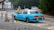 A light blue taxi is parked on the side of a road, near a sign that reads 'Alighting Only'. The vehicle has a yellow license plate with the number KY 8292, and the word 'TAXI' is displayed on its roof. There are several road signs and a small grey car in the background, indicating a quiet urban setting with surrounding greenery.