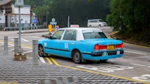 A light blue taxi is parked on the side of a road, near a sign that reads 'Alighting Only'. The vehicle has a yellow license plate with the number KY 8292, and the word 'TAXI' is displayed on its roof. There are several road signs and a small grey car in the background, indicating a quiet urban setting with surrounding greenery.