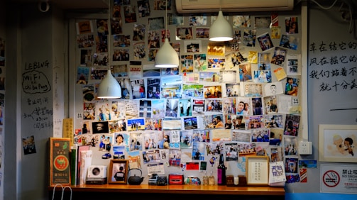 A wall filled with a collage of various photographs and handwritten notes, illuminated by three hanging pendant lights. The photos include people, places, and text in different languages. The bottom area has a wooden ledge with books, decorative items, and a few small framed pictures.